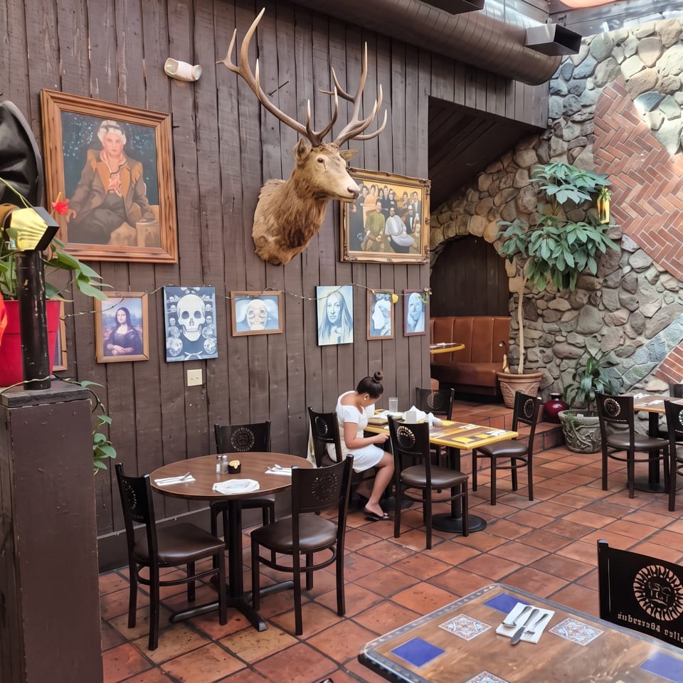 Rustic restaurant interior with tiled floors, wooden walls, framed artwork, a mounted deer head, stone fireplace, and dining tables.