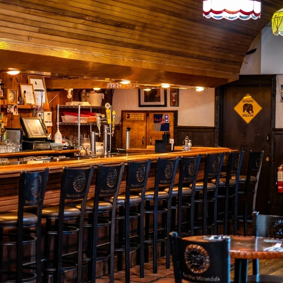 Cozy bar interior with a long wooden bar, stools, liquor bottles, and small round tables.