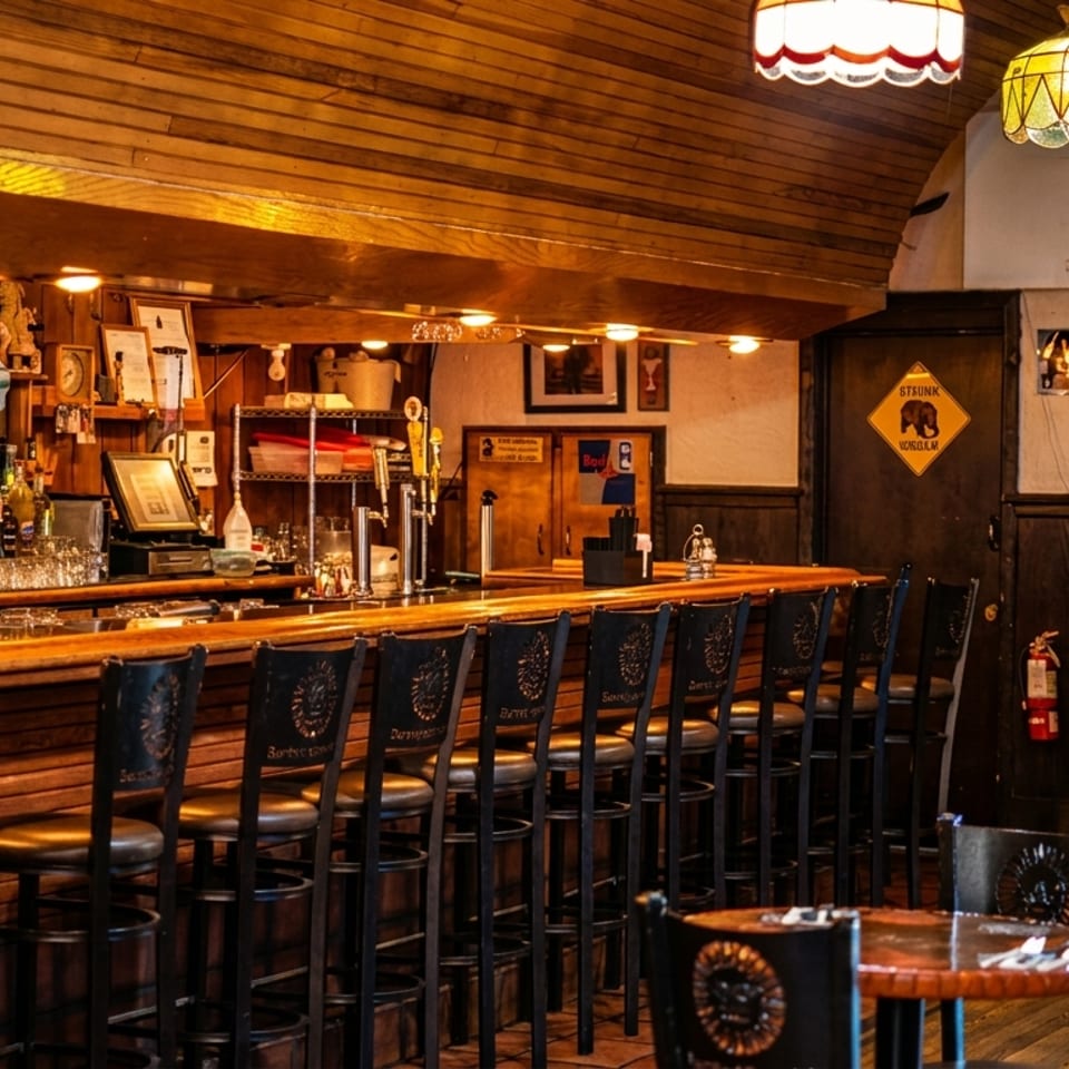 Cozy, dimly lit bar interior with a long wooden counter, bar stools, and various liquor bottles.