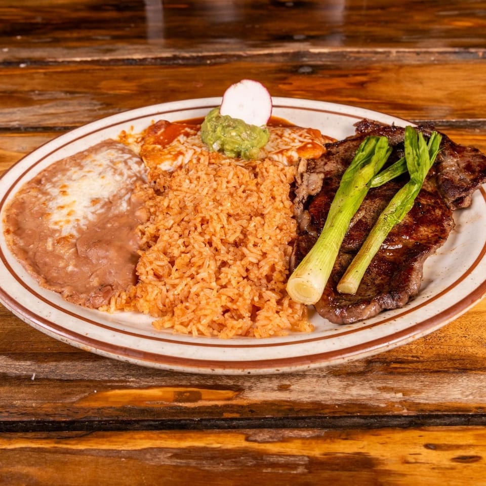 Mexican-style platter with grilled steak, rice, refried beans, enchiladas, guacamole, radish, and grilled green onions on a wooden table.