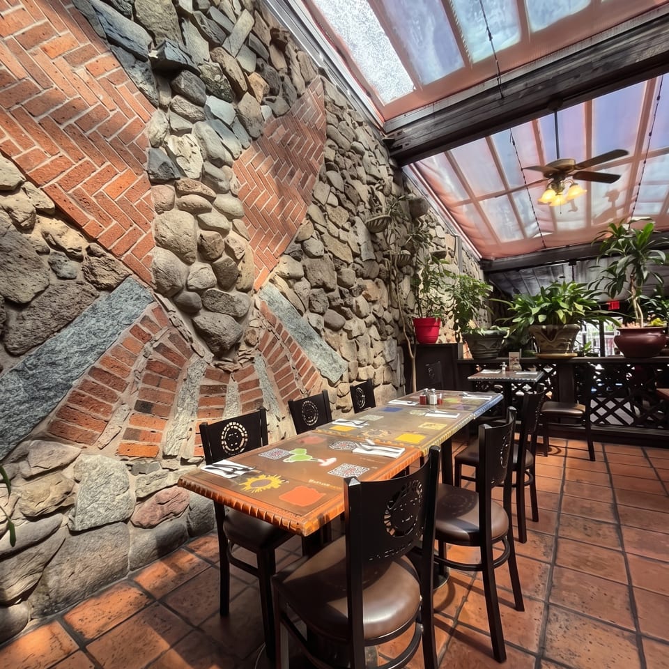 Restaurant dining area with stone and brick walls, set tables, potted plants, and a bright glass-paneled ceiling.