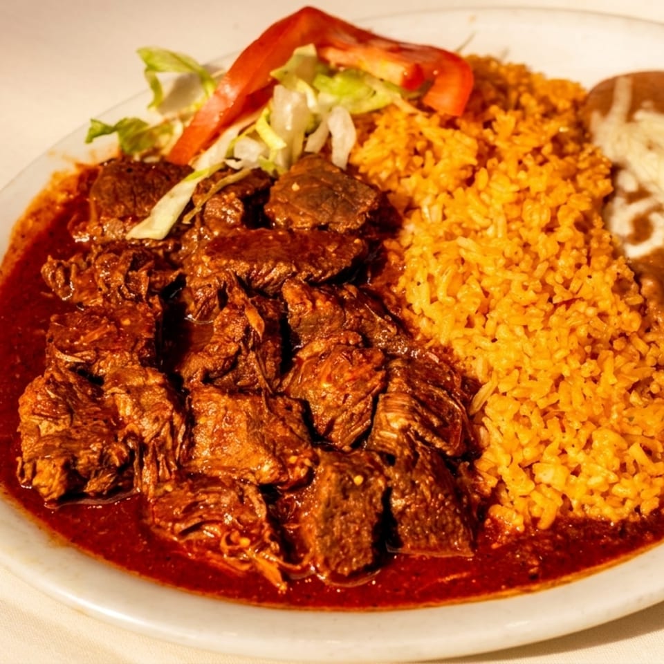 Plate of carne guisada with red rice, refried beans, shredded cheese, lettuce, and tomato.