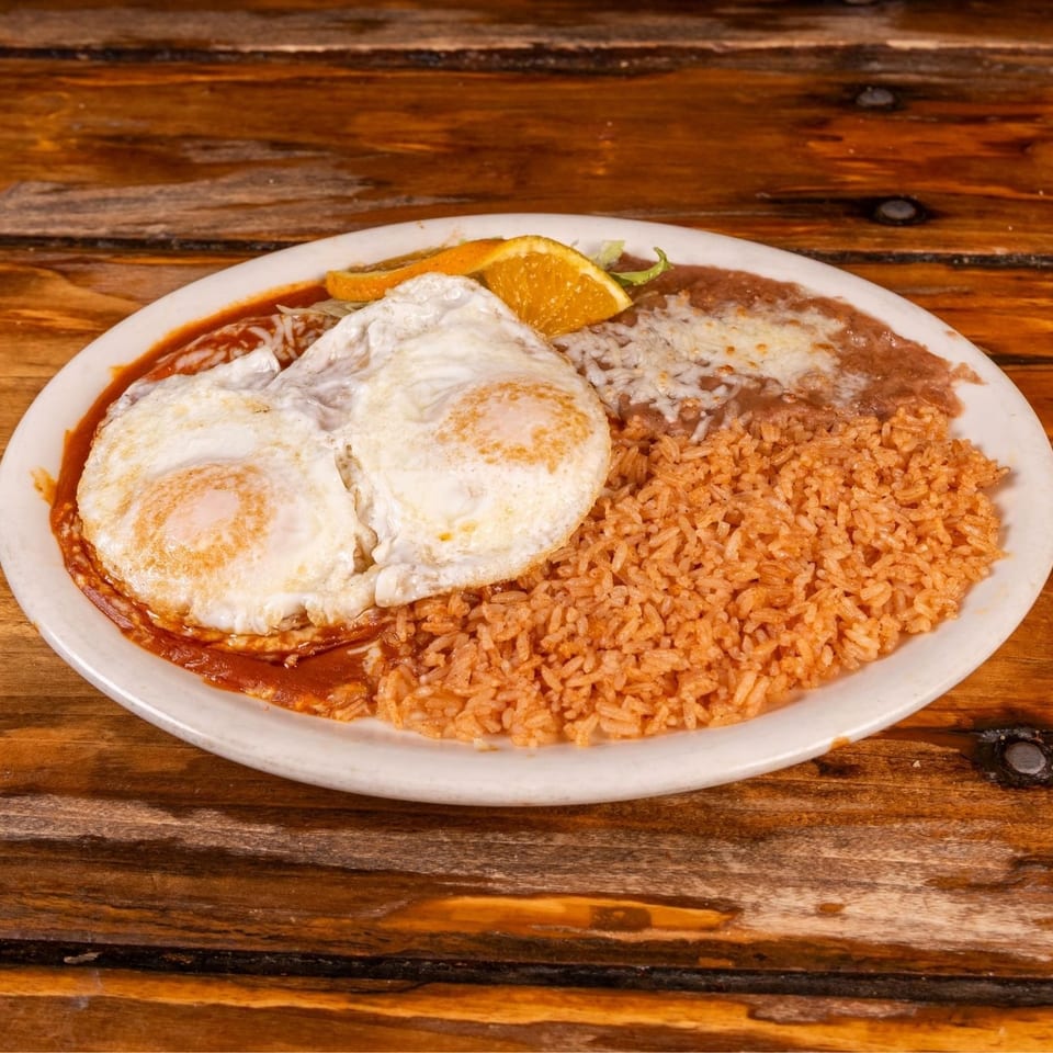 Plate of Mexican-style rice, refried beans, red sauce, and two fried eggs garnished with orange slices on a wooden table.