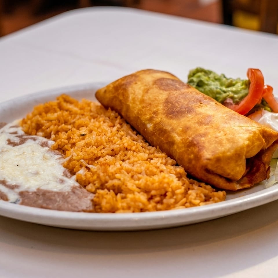 Golden-fried chimichanga with rice, refried beans, guacamole, sour cream, and diced tomatoes on a white plate.