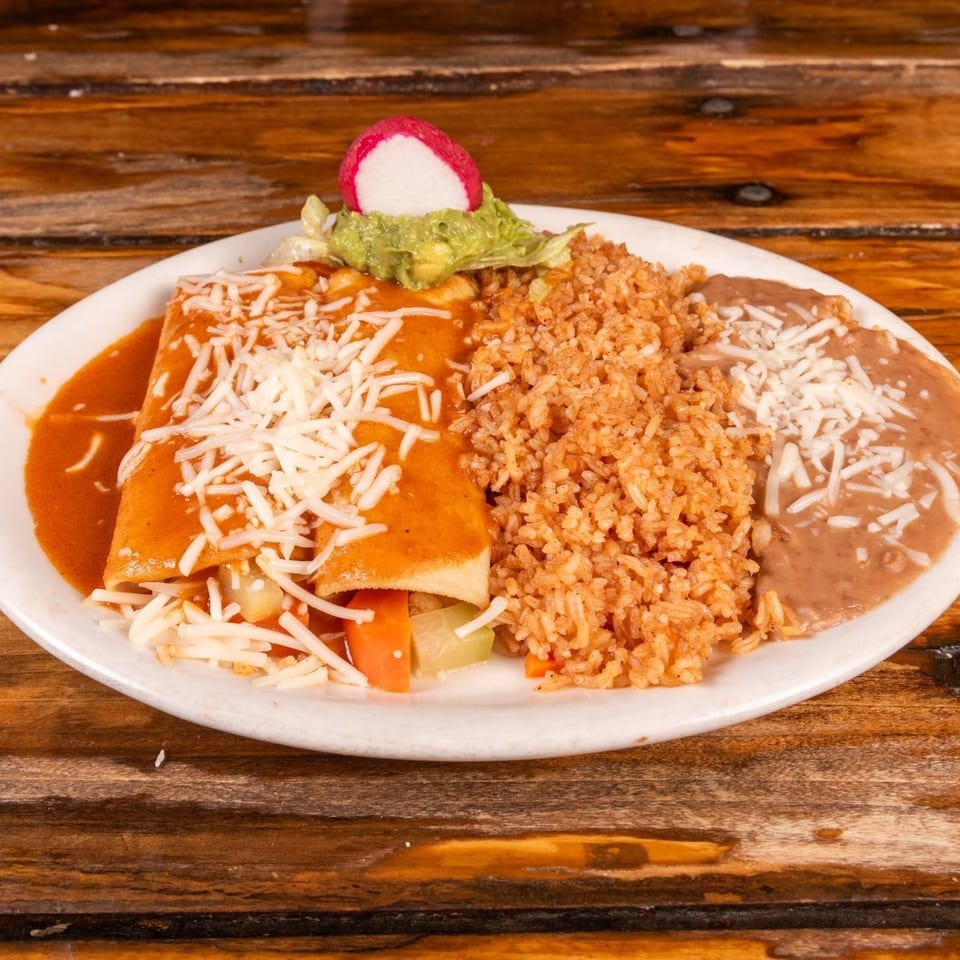 Mexican plate featuring cheese-covered enchiladas, Spanish rice, refried beans, guacamole, radish, lettuce, and vegetables on a wooden table.